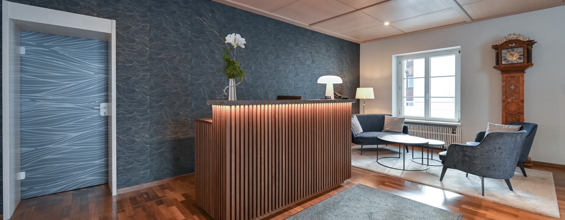 Rezeption vom Gästehaus Sankt Ulrich mit beleuchteter Holztheke, blauer Wand mit Muster, zwei blauen Sesseln, Couchtisch, Stehlampe und antiker Standuhr neben Fenster.