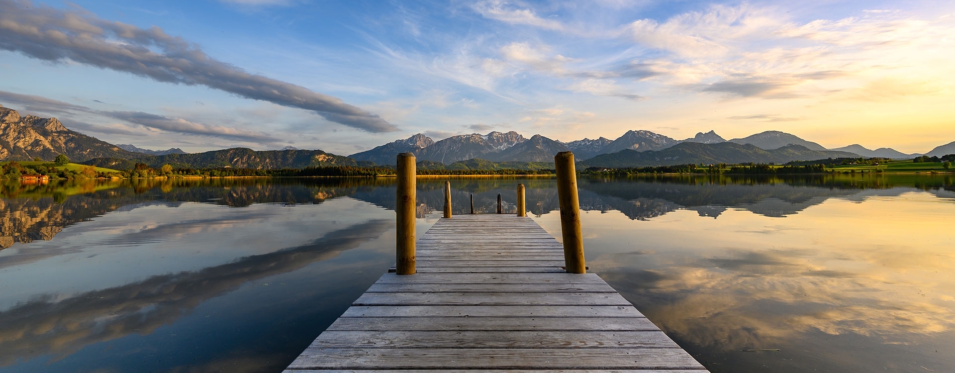 Holzsteg führt auf den ruhigen Hopfensee mit Spiegelung von Bergen und bewölktem Himmel bei Sonnenuntergang.