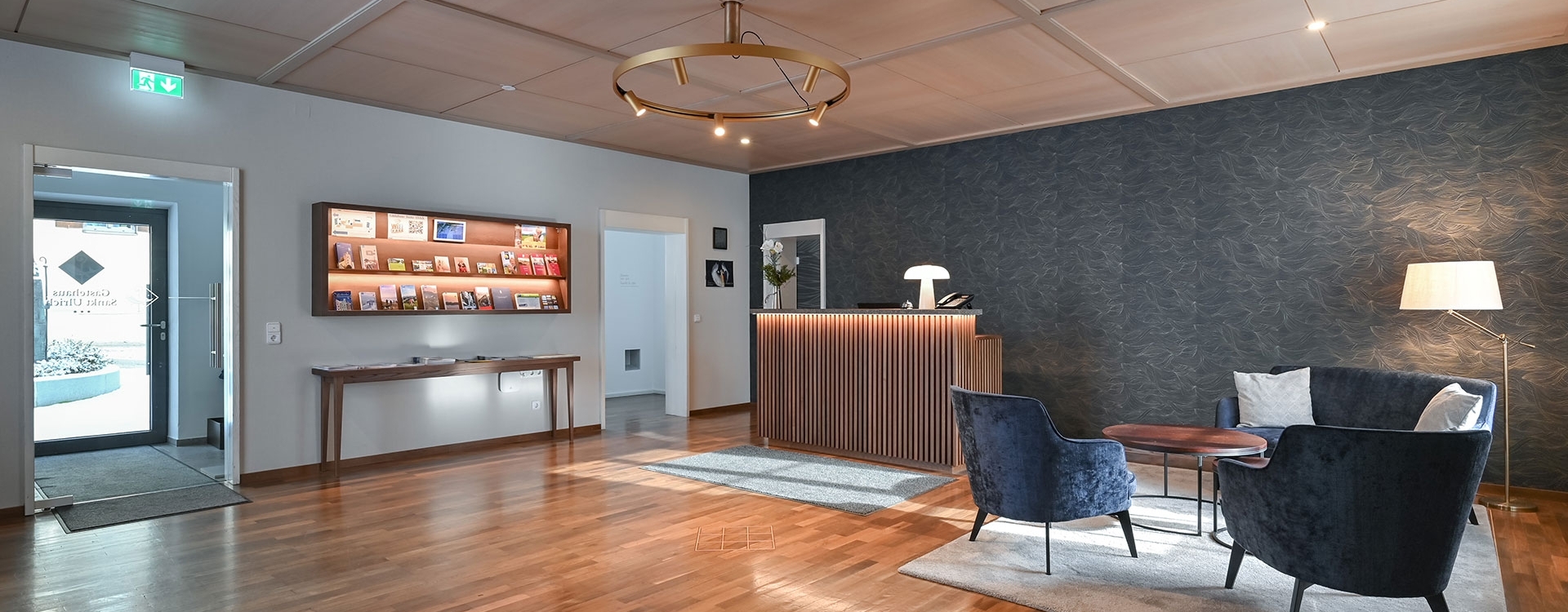Moderner Empfangsbereich vom Gästehaus Sankt Ulrich mit Holztresen, beleuchtetem Prospektregal, grauer Couch mit weißen Dekokissen und Stehlampe.