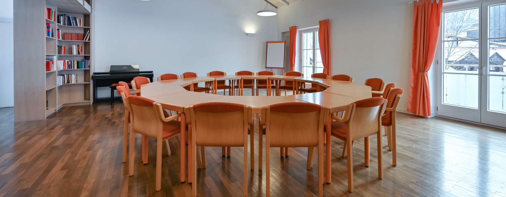 Konferenzraum im Gästehaus Sankt Ulrich mit rundem Tisch, orangefarbenen Stühlen, Holzboden, Bücherregal, Klavier und großen Fenstern mit orangen Vorhängen.