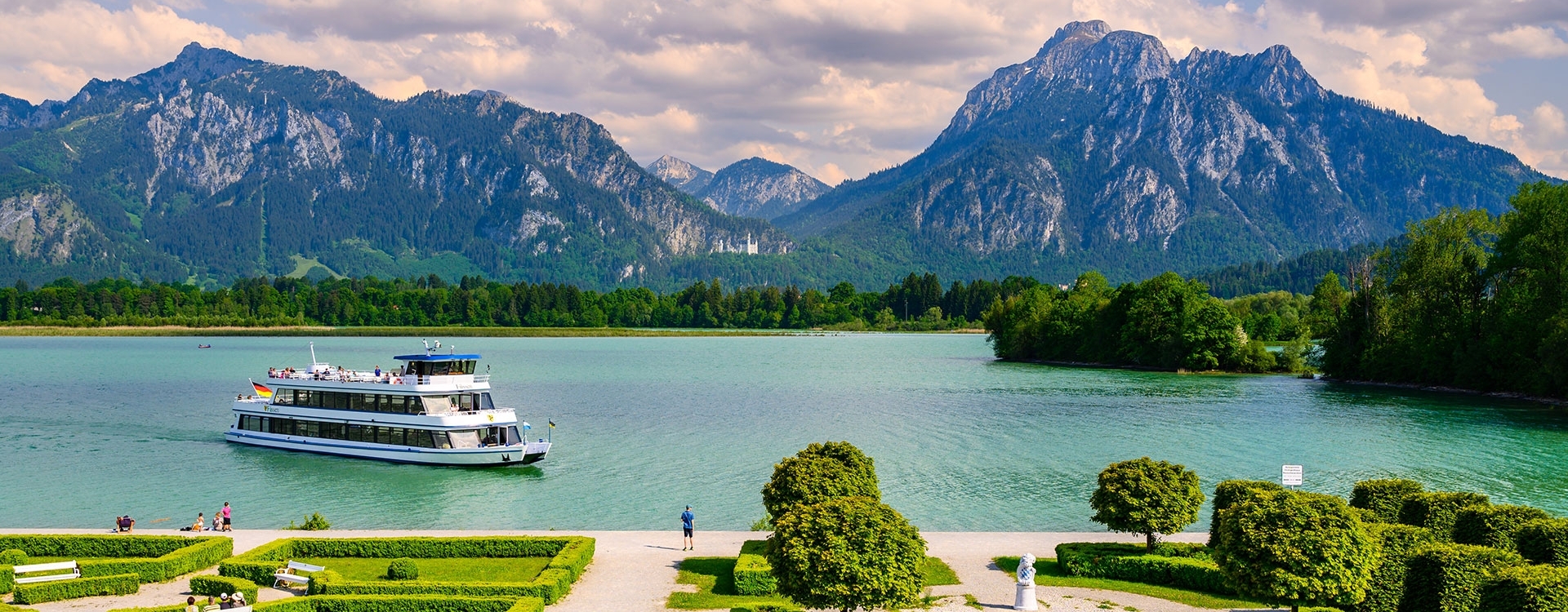 Schiff MS Füssen auf dem Forggensee mit Alpen im Hintergrund, links der Tegelberg und rechts der Säuling mit bewaldetem Ufer und bewölktem Himmel.