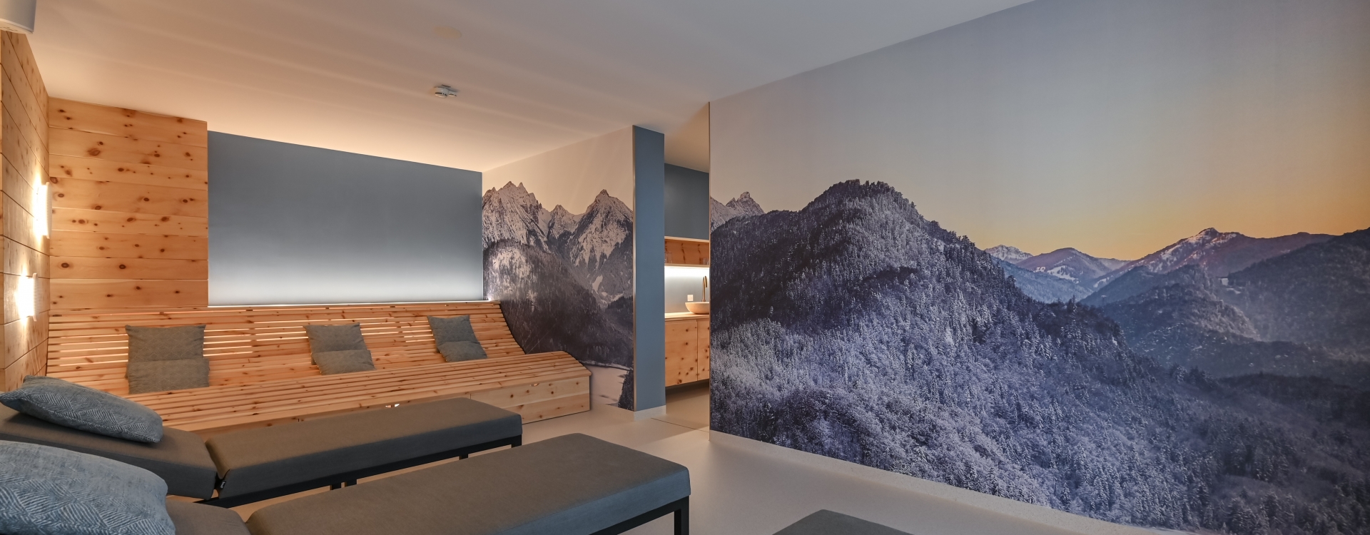 Innenraum des Ruhebereiches im Gästehaus St. Ulrich mit Holzverkleidung, Sitzbank mit grauen Kissen und Wandbild von verschneiten Bergen bei Sonnenuntergang.