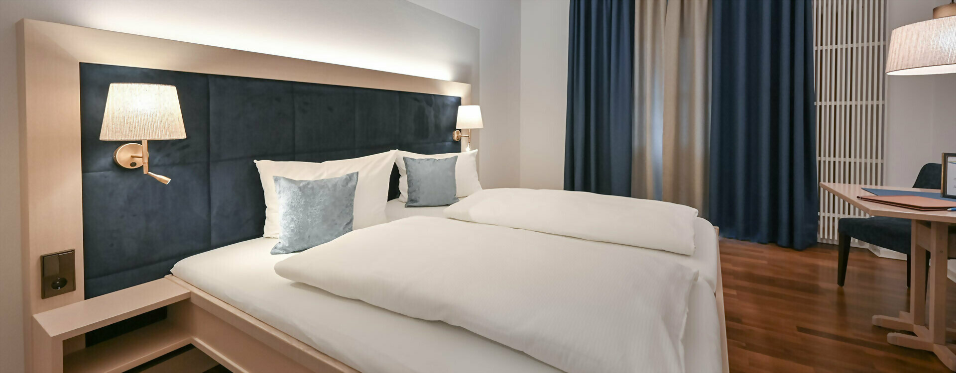 Zimmer mit Doppelbett im Gästehaus Sankt Ulrich, dunkelblauem gepolstertem Kopfteil, weißen Bettdecken und grauen Kissen, Wandlampen und dunklen Vorhängen.