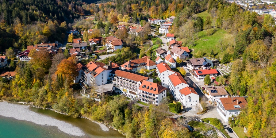 Luftaufnahme von Bad Faulenbach mit Häusern mit roten Dächern, umgeben von herbstlich gefärbtem Wald und dem Flussufer vom Lech.