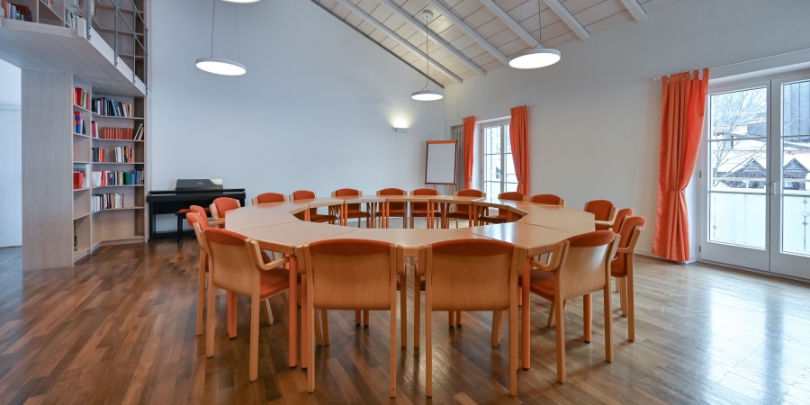 Konferenzraum mit rundem Tisch, orangefarbenen Stühlen, Holzboden, Bücherregal, Klavier und großen Fenstern mit orangen Vorhängen.