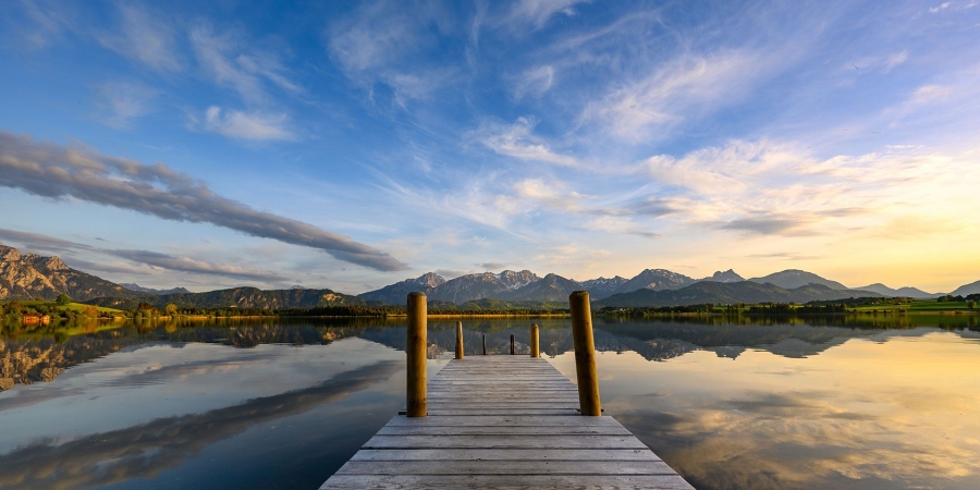 Holzsteg führt auf den ruhigen Hopfensee mit Spiegelung von Bergen und bewölktem Himmel bei Sonnenuntergang.