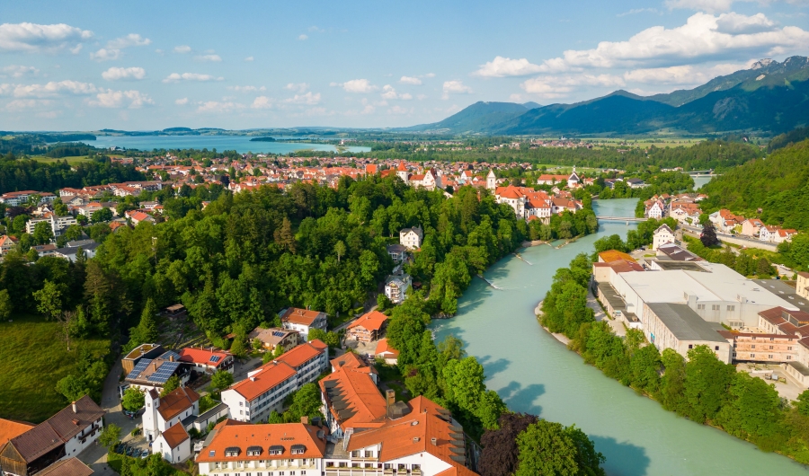 Luftaufnahme von Bad Faulenbach und Füssen mit roten Dächern, dem Fluss Lech, der sich durch die Stadt schlängelt, grünen Wäldern und den Alpen im Hintergrund unter blauem Himmel.