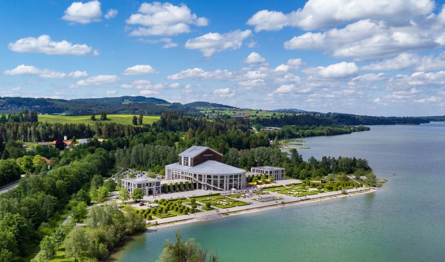 Festspielhaus Füssen mit Gartenanlagen direkt am Ufer des Forggensees, umgeben von Wald und Feldern unter blauem Himmel mit Wolken.