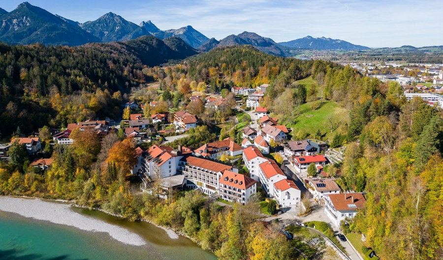 Luftaufnahme von Bad Faulenbach und Füssen mit roten Dächern, dem Fluss Lech, der sich durch die Stadt schlängelt, grünen Wäldern und den Alpen im Hintergrund unter blauem Himmel.