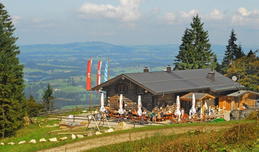 Berggasthaus ‚Drehhütte‘ mit Holzbau, Sonnenschirmen und Gästen auf einer Terrasse, umgeben von Wiesen und Tannenbäumen, mit Blick auf ein Tal und Berge im Hintergrund.