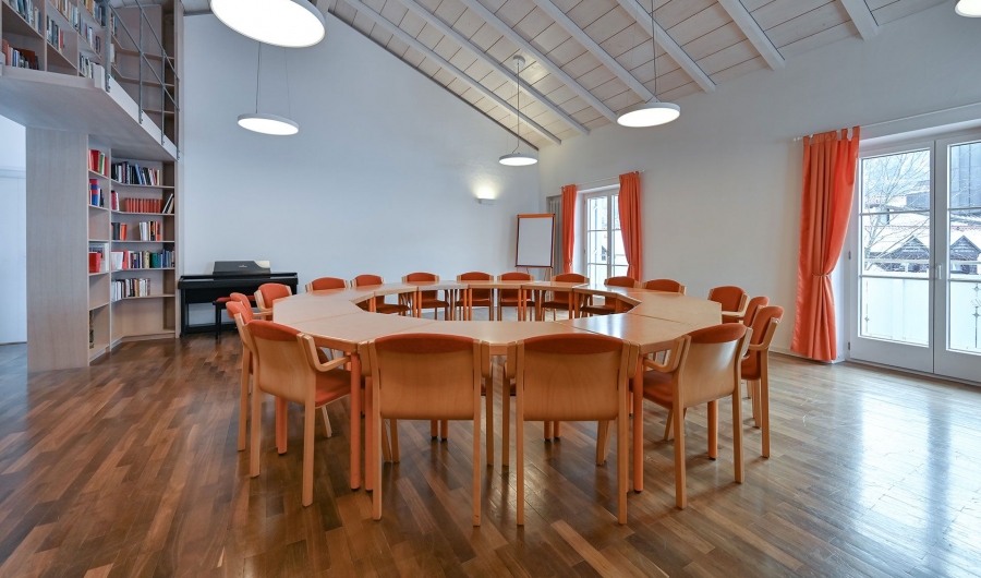Seminarraum im Gästehaus Sankt Ulrich mit rundem Tisch, orangefarbenen Stühlen, Holzboden, Bücherregal, Klavier und großen Fenstern mit orangen Vorhängen.