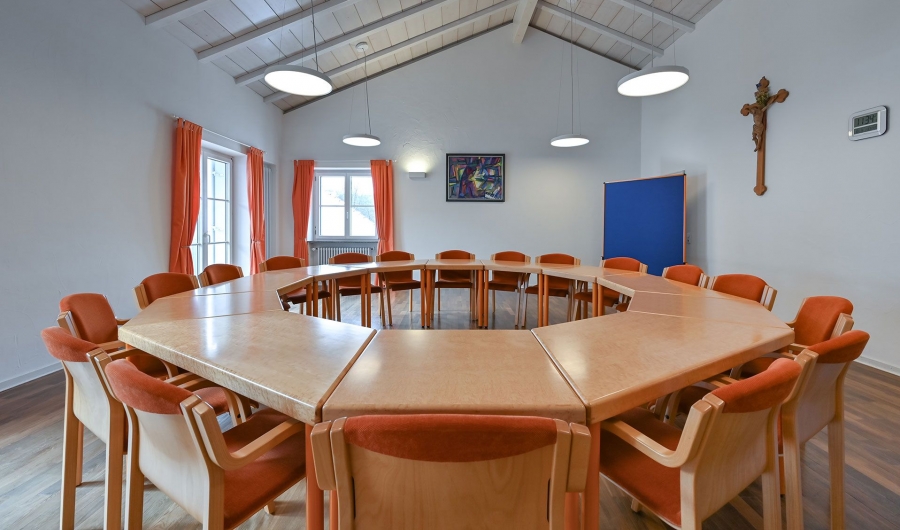 Konferenzraum im Gästehaus Sankt Ulrich mit zu einem Kreis aufgestellten Tischen und orangefarbenen Stühlen, weiße Wände, zwei Fenster mit orangen Vorhängen, Kreuz an der Wand.