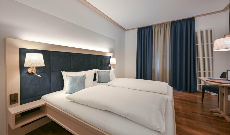 Modernes Hotelzimmer im Gästehaus Sankt Ulrich mit großem Doppelbett, dunkelblauem gepolstertem Kopfteil, weißen Bettdecken und Kissen, Holzboden und dunkelblauen Vorhängen.