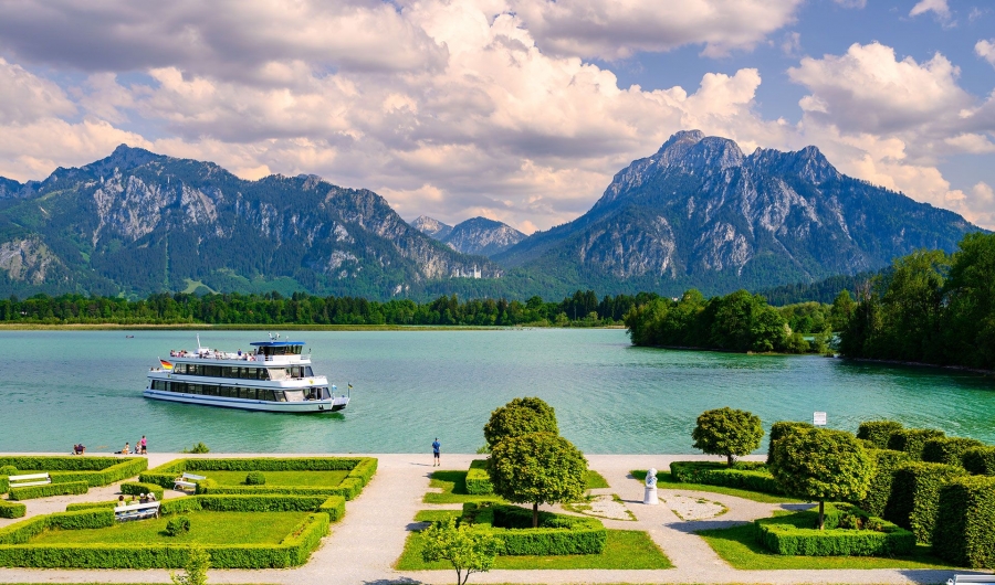 Das Schiff MS Füssen fährt auf dem Forggensee vor dem Festspielhaus Füssen mit den Alpen und bewölktem Himmel im Hintergrund.
